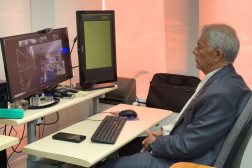 Foto mostra um homem sentado em uma cadeira, diante de um computador. Ele participa de uma reunião online. 