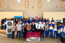 Foto mostra várias pessoas reunidas em um salão. Todos estão juntos em pé. São crianças, adolescentes e pessoas adultas reunidas.