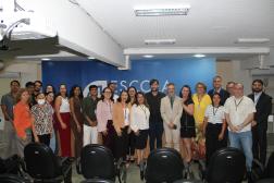 Foto mostra várias pessoas em pé, dentro de um auditório. Ao fundo, parede azul.
