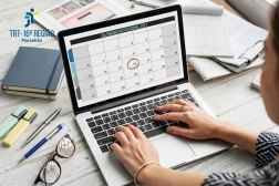 Foto de uma mesa de trabalho com mãos femininas no teclado de um notebook, em cuja tela aparece um calendário.