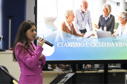 Foto mostra uma mulher usando um blaser na cor rosa. Ao fundo um telão projeto uma imagem com idosos reunidos. E a mensagem etarismo e ciclo da vida
