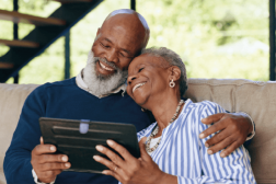 Foto de um homem e mulher negros e idosos abraçados e sorrindo, olhando a tela de um tablet, sentados em um sofá.