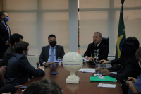 Fundo branco com imagem do presidente do TRT, desembargador Carvalho Neto, trajando terno escuro, estando sentado ao lado do procurador do trabalho Maurício Pessoa Lima, vestido com terno escuro e camisa e gravata azuis. Ambos estão de frente para os demais participantes da audiência que se encontram sentados em torno da mesa de reunião.