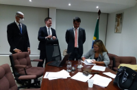 Foto da desembargadora Márcia Andrea, sentada à mesa de reunião no Gabinete da Presidência, vestindo blazer cinza, usando óculos, assinando ata de audiência de conciliação, observada pelo procurador do trabalho Roberto Moreira, pelo assessor do gabinete da desembargadora, Hermano Rocha Filho, e o procurador do município Bruno Duailibe, os três em pé, com ternos escuros