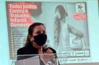 Imagem da juíza Liliana Bouéres, vestindo casaco preto, usando máscara preta, com cabelo preso e segurando microfone, tendo ao fundo o cartaz em tela com uma menina de costas engomando roupas e texto informativo Todos Juntos Contra o Trabalho Infantil Doméstico na cor branca sobre fundo vermelho.