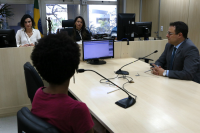 Imagem de uma sala de audiência onde estão três mulheres e um homem. No primeiro plano, uma mulher negra de costas, vestindo blusa na cor vinho, em frente de um homem branco, cabelos pretos, usando óculos, vestindo terno cinza, sentados junto a uma mesa de cor bege, tendo á frente uma tela de computador e microfones. No segundo plano, uma mulher de cabelos pretos acima dos ombros e vestindo blusa branca, ao lado de outra mulher de cabelos longos, lisos, vestindo uma toga