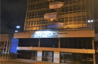 Foto da fachada do prédio-sede do TRT no Maranhão, no bairro Areinha. A fachada está iluminada na cor amarela em adesão ao Maio Amarelo, e se vê partes do projeto arquitetônico nas cores azul e bege, uma pequena grande antes do acesso à Portaria do Tribunal (frontal), na lateral esquerda os mastros de hastear as bandeiras oficiais