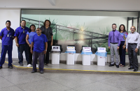 Foto de servidores, servidoras, terceirizadas e terceirizados em um dos ambientes do prédio-sede do Tribunal do Trabalho onde se vê de coletores de papel, plástico, metal e lixo comum, ao fundo uma parede de vidro e parte de parede pintada na cor branca