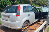 Foto de um carro da Ford cinza, com detalhes nas luminárias na cor vermelha, com a porta de passageiro aberta, colocado sobre um guincho. ao fundo uma área verde com várias árvores