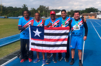 Foto de atletas do TRT do Maranhão com o treinador Emerson Viana. A foto foi feita numa pista de atletismo, localizada numa área verde. Os atletas e o técnico estão com camisas azuis e seguram a bandeira do Maranhão. Ao fundo, há um tenda branca, carros estacionados e uma casa.