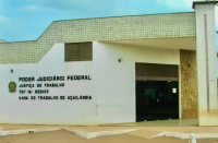 Foto da fachada principal da Vara do Trabalho de Açailândia, localizada no Sul do Maranhão, no primeiro plano uma parede branca com o brasão do Poder Judiciário e ao lado está escrito o Poder Judiciário Federal, Justiça do Trabalho, TRT-16ª Região, Vara do Trabalho de Açailândia, ao lado uma porta de vidro escura e demais partes da fachada na cor bege com tons de caramelo