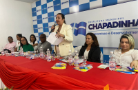 Imagem da juíza Liliana Bouéres proferindo discurso em pé com microfone ao centro da mesa do evento composta por autoridades que se encontram sentadas. A mesa está coberta por uma toalha vermelha e ao fundo tem um painel em tons branco e azul.