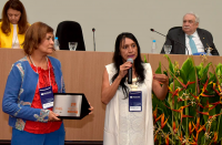 Foto das servidoras do TRT do Maranhão Edvânia Kátia (roupa clara), usando crachá azul e discursando após a conquista do prêmio na categoria especial (ramo Justiça do Trabalho) e Nonata Teixeira (roupa azul e vermelha), que está segurando o prêmio; ao lado um arranjo de flores; as servidoras estão em frente à mão de honra do evento