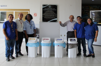 Foto de servidores, terceirizadas e terceirizados no saguão do Fórum Astolfo Serra onde foram disponibilizados coletores de papel, plástico, metal e lixo comum, na frente de uma parede branca onde está fixada uma placa inauguração do Fórum