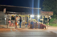 Imagem noturna de banca de frutas em rodovia onde se vê três pessoas.