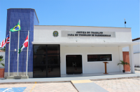 Foto da fachada principal da Vara do Trabalho de Barreirinhas, no Estado do Maranhão. No primeiro plano, entrada da VT com rampa e porta de vidro escuro. Paredes brancas e janelas de vidro escuro. À esquerda, coluna em lajotas azuis, à frente da qual veem-se as bandeiras do Brasil (centro), do Maranhão (esquerda) e do Município (direita). No topo, brasão do Poder Judiciário e, ao lado deste, as inscrições Justiça do Trabalho e Vara do Trabalho de Barreirinhas.