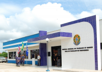 Foto da fachada principal da sede da Vara do Trabalho de Chapadinha. A fachada pintada nas cores azul celeste e branca, com detalhes em cerâmica azul marinho. Portas e janelas de vidro transparente. Na frente da fachada, policiais e um vigilantes e mais duas pessoas. Também estão hasteadas as bandeiras do Brasil, Maranhã e de Chapadinha. Na parede principal da fachada há um brasão e abaixo está escrito: Tribunal Regional do Trabalho da 16ª Região - Vara do Trabalho de Chapadinha