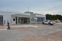 Foto da fachada principal da sede da Vara do Trabalho de São João dos Patos. A fachada está pintada na cor branca. A sede tem portas de vidro transparente e persianas nas janelas. Próximo à porta, dois vasos com plantas, e à esquerda  da porta estão hasteadas as bandeiras do Brasil, Maranhão de São João dos Patos. Na parede, à direita, há um brasão e abaixo está escrito: Justiça do Trabalho - Vara do Trabalho de São João dos Patos. No estacionamento, há um veículo oficial do TRT e uma moto.