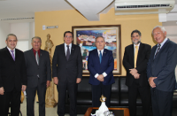 Cinco desembargadores do TRT e o ministro do TST em pé, lado a lado, posando para o registro fotográfico. Ao fundo, parede amarela, quadro com imagens de casas em espaço urbano.