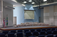 Foto com vista panorâmica do palco do Auditório Ari Rocha com os alimentos a serem doados dispostos ao centro. Bandeiras do Maranhão e do Brasil hasteadas à esquerda. Cadeiras azuis do Auditório na parte de baixo da foto. Telão de projeção de imagens no topo, sobre parede azul claro.