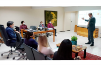 Foto no Gabinete do corregedor regional, desembargador Gerson de Oliveira, durante apresentação ao corregedor-geral da Justiça do Trabalho, ministro Caputo Bastos, equipe da Corregedoria. O desembargador Gerson está em pé, e o ministro e demais pessoas sentadas em cadeiras na cor cinza.  Sala na cor bege, onde se vê bancada e mesa de centro de madeira, mesa grande de madeira com tampo de vidro, um quadro pendurado na parede,