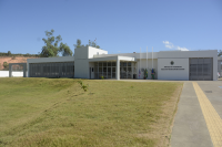 Foto da sede da Vara do Trabalho de Caxias. No primeiro plano, vê-se o gramado. Ao fundo, a fachada principal da sede da VT pintada na cor branca. À direita, mastros com as bandeiras do Brasil, Maranhão e Caxias. Ao lado, uma parede branca com o brasão do Poder Judiciário e abaixo está escrito: Justiça do Trabalho, Vara do Trabalho de Caxias. Portas de vidros e portas e janelas com persianas claras. Por trás da sede, vê-se copas de arvores.