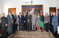 Treze autoridades de pé em semicírculo, em sala de gabinete com tapete, tendo ao fundo parede branca e grande quadro horizontal com a deusa da Justiça. 