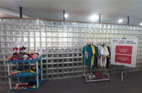 Foto da entrada do Auditório com parede de tijolos de vidro ao fundo. À esquerda, estante baixa com as bonequinhas de pano da Casa Acolher. À direita, arara de roupas com camisetas e canecas, e banner do Pouso Obras Sociais.