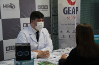 Foto de um homem e uma mulher: ele, médico oftalmologista, usando jaleco branco por cima de camisa branca com alguns detalhes e gravata preta; ela usando um blazer preto. Atrás, na parede, logomarcas do HRO e Geap.