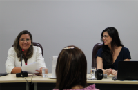 Foto de duas mulheres sentadas à mesa de honra do evento. As duas têm cabelos lisos, compridos e usam óculos. A da esquerda está usando blazer e a da direita está usando uma roupa azul marinho.