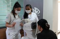 Foto de três mulheres: uma técnica de enfermagem e uma atendente do HRO, ambas vestidas com roupas brancas, e de uma terceirizada usando blazer preta, que está sentada diante de um aparelho usado para fazer exames oftalmológicos