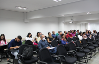 Foto de várias pessoas (mulheres e homens) sentados em cadeiras pretas, assistindo aula num auditório