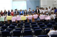 Foto de diversas pessoas (adultos e jovens) na frente e sobre o palco do auditório onde ocorreu a audiência pública