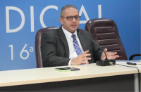 Foto do desembargador James Magno, usando óculos, terno escuro, camisa branca e gravata azul, gesticulando com ambas as mãos, sentado à mesa, falando ao microfone. Ao fundo, parede azul com parte de inscrição branca.