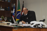 Foto com fundo branco e bandeiras do Maranhão, Brasil e da Justiça do Trabalho, tendo ao centro o presidente do TRT no Maranhão, desembargador Carvalho Neto, sentado em seu gabinete, vestindo terno escuro.