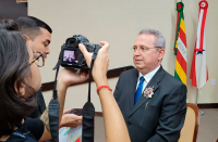 Desembargador Carvalho Neto portando medalha no terno escuro. Encontra-se em posição de registro fotográfico para duas pessoas, uma utiliza câmera fotográfica e a outra celular. Ao fundo duas bandeiras em mastros.