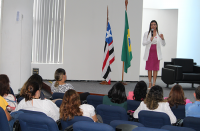 Foto de pessoas sentadas e de uma mulher em pé no palco de um auditório, próximo às bandeiras do Brasil e do Maranhão e de uma poltrona preta.