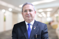 Foto do desembargador Carvalho Neto, usando terno azul escrito, camisa branca e gravata azul roial, com fundo desfocado claro, em tons de bege.