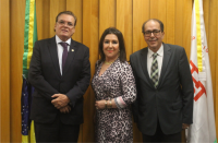 Foto de dois homens e uma mulher, em pé, à frente de parede amadeirada. Atrás estão duas bandeiras.