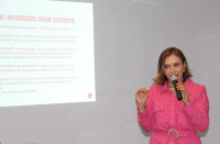 Foto de uma mulher que está em pé, falando ao microfone, vestindo roupa na cor rosa