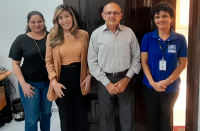 Foto de três mulheres e um homem que estão em pé, em frente a uma porta de madeira, numa sala onde se vê uma mesa, computadores e outros materiais.