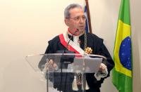 Foto de um homem usando toga, na tribuna. Ao fundo, bandeira do Brasil .