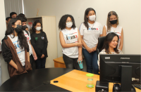 Foto de estudantes em pé, usando máscaras, observando tela de computador,  próximos a uma mulher de cabelos longos e escuros, sentada.