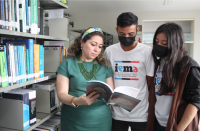 Foto de um casal de um rapaz e uma moça, observando um livro mostrado por uma mulher de cabelos claros, usando vestido e tiara verdes.