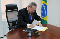 homem de terno sentado atrás da mesa assinando documento com bandeira do Brasil ao fundo.