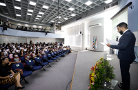 Foto de um homem em pé em um palco falando ao microfone e de uma plateia