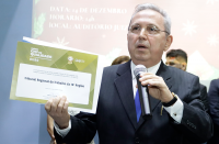 homem de terno, com microfone em um mão e na outra um certificado 