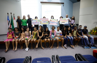 Foto de adolescentes, jovens e adultos (homens e mulheres), alguns sentados e outros em pé em um palco