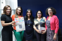 cinco mulheres posam para foto, uma das quais segura um cartaz do inovalab. Ao fundo, parede azul e janela com iluminação solar.