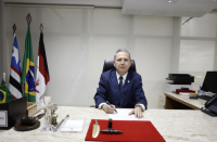 homem de terno sentado à mesa de gabinete. Ao fundo, bandeiras hasteadas. 